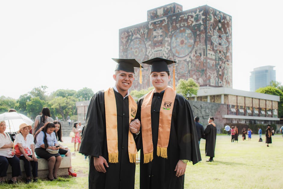 Two graduates posing in front of a university building during a sunny graduation ceremony.