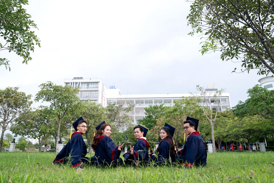Group of five university graduates celebrating outdoors in regalia, sitting with a campus backdrop.